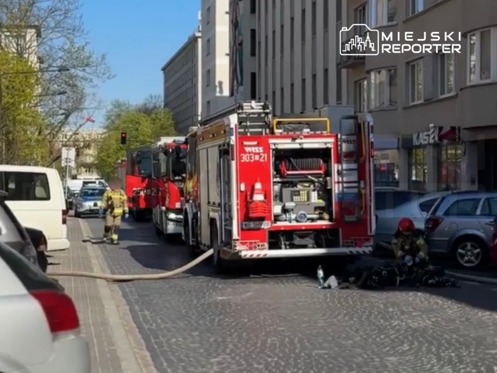 Strażak w mundurze z węża gaśniczego obok wozu strażackiego na ulicy, obok zaparkowane samochody.