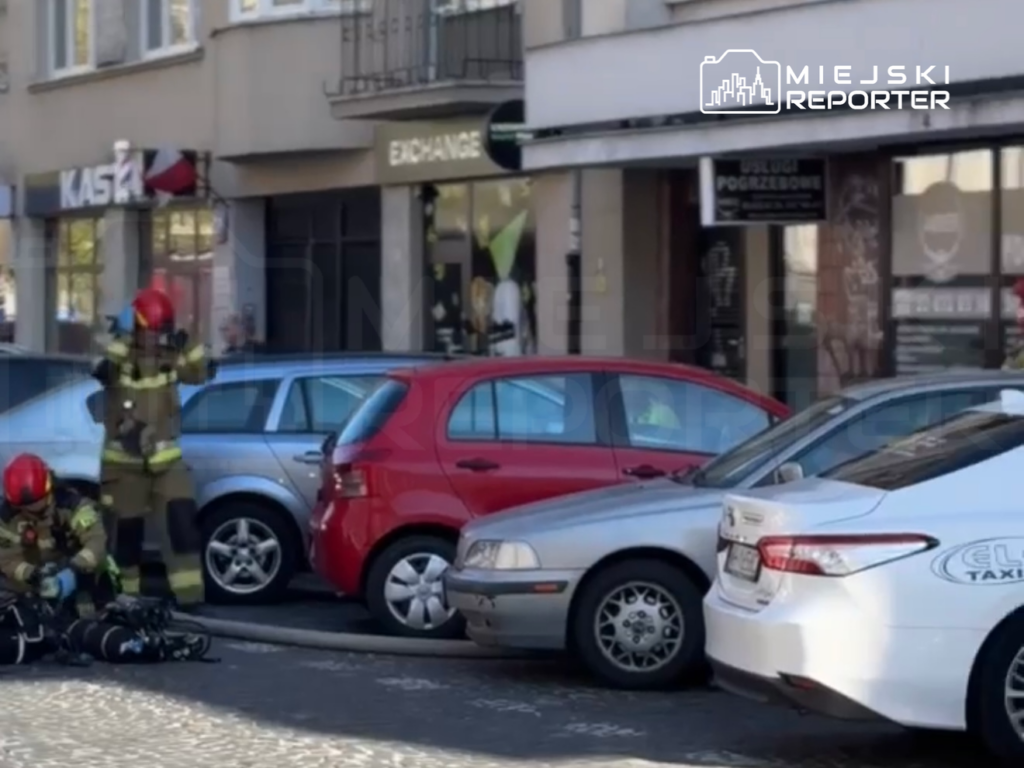 Strażacy w hełmach i mundurach przygotowują sprzęt na chodniku obok zaparkowanych samochodów.