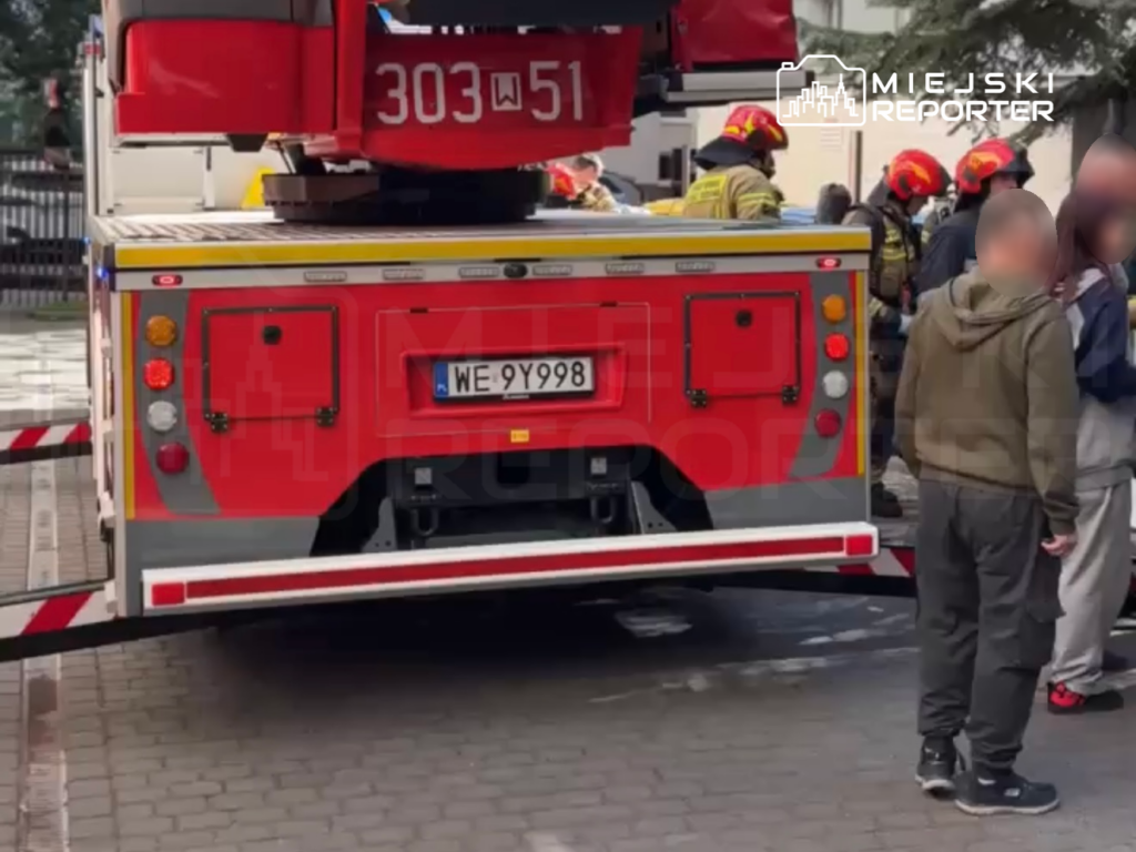 Strażacy w mundurach omawiają sytuację obok wozu strażackiego na ulicy, w tle widoczne są osoby cywilne.