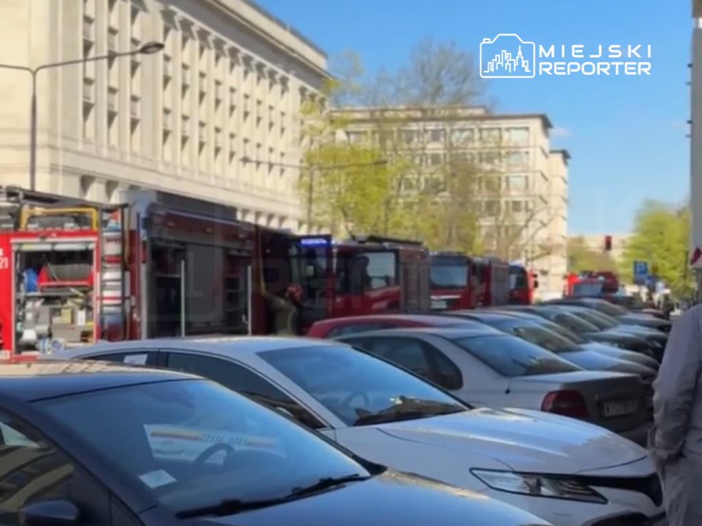 Czerwone wozy strażackie zaparkowane przy budynku, obok rzędy zaparkowanych samochodów osobowych.