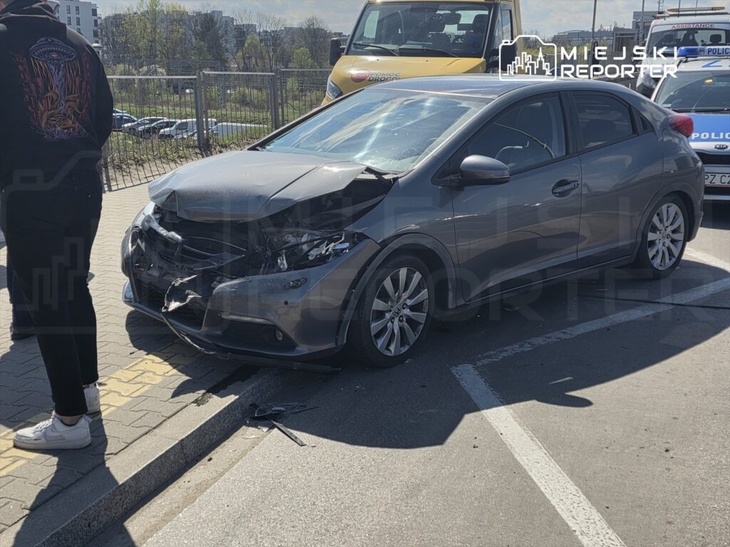 Zniszczony samochód osobowy stoi na parkingu, obok mężczyzna w czarnej bluzie obserwuje sytuację.