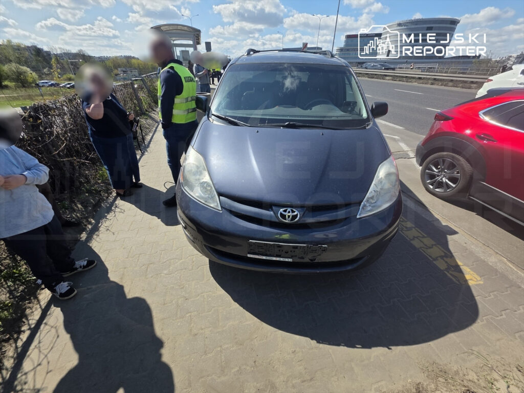 Mężczyzna w kamizelce odblaskowej rozmawia z kobietą obok zaparkowanego samochodu na chodniku przy ruchliwej drodze.