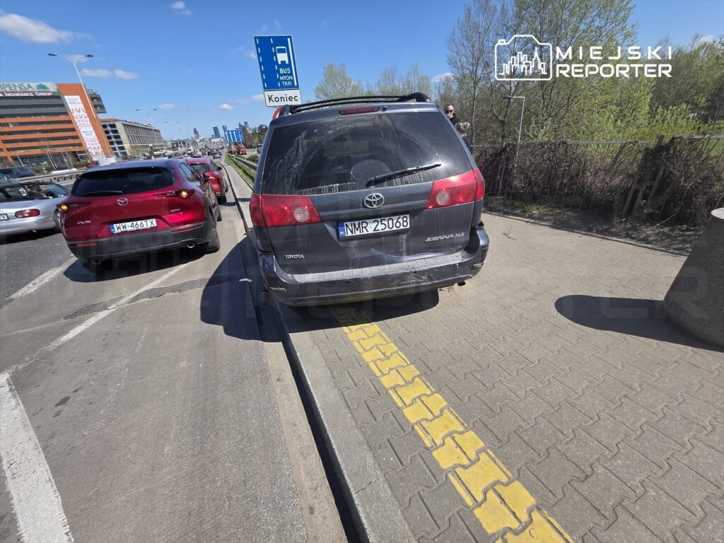 Czerwony samochód osobowy zatrzymany obok szarego SUV-a na ruchliwej ulicy, w tle widoczne biurowce.