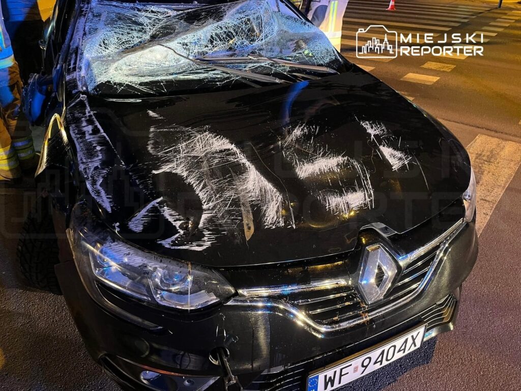 Uszkodzony czarny samochód osobowy z rozbitym przednim szybą i widocznymi rysami na karoserii, stojący na ulicy.