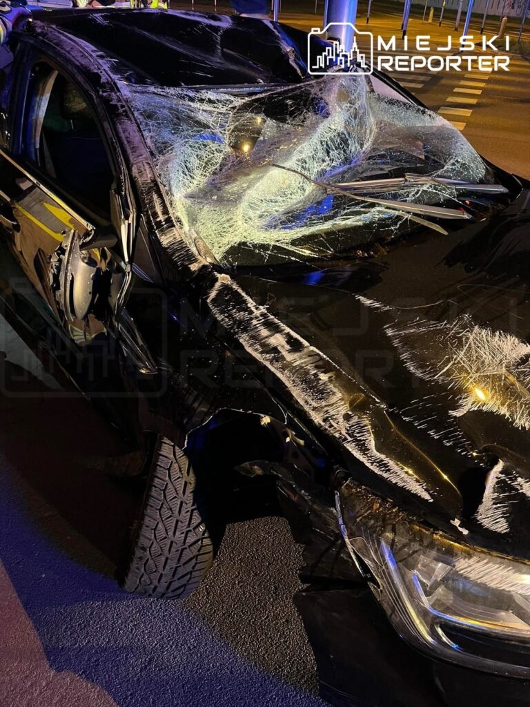 Zniszczony czarny samochód osobowy z rozbitym przednią szybą, zaparkowany na ulicy w nocy.