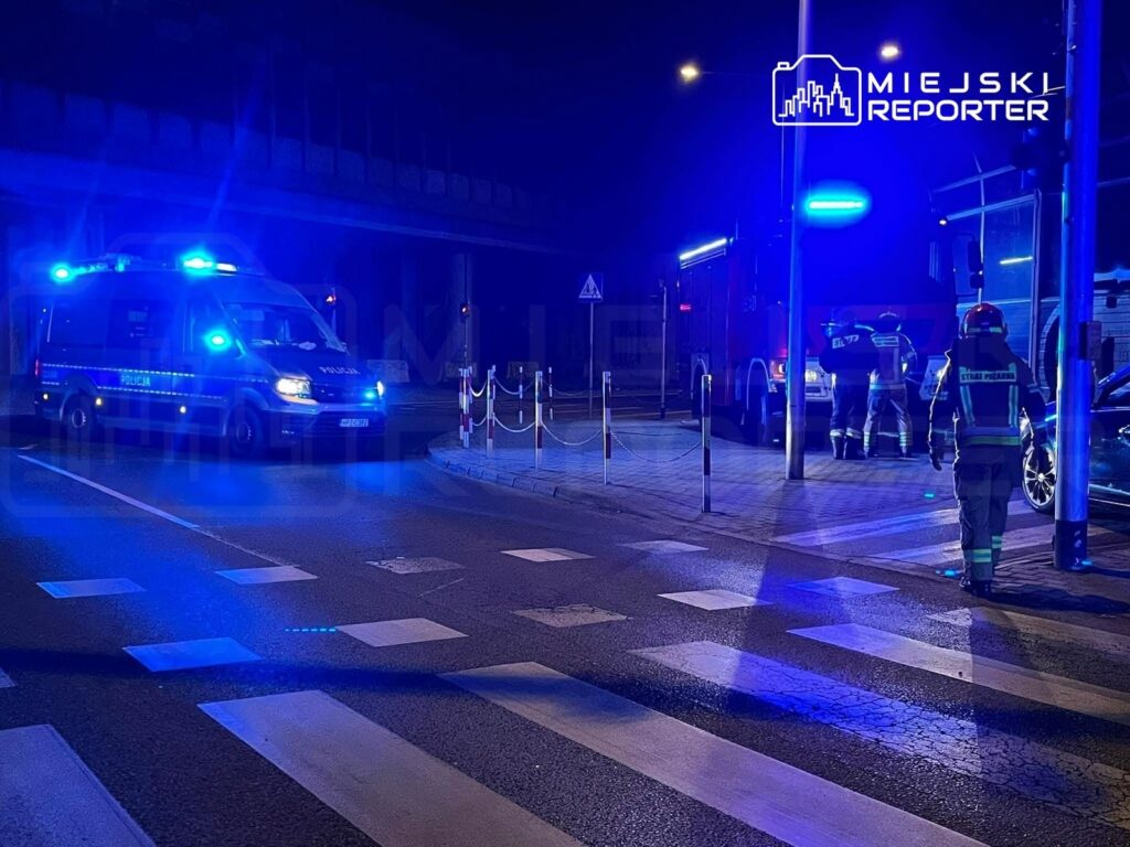 Radiowóz policyjny z włączonymi sygnałami świetlnymi stoi na skrzyżowaniu, a obok niego pracują strażacy.