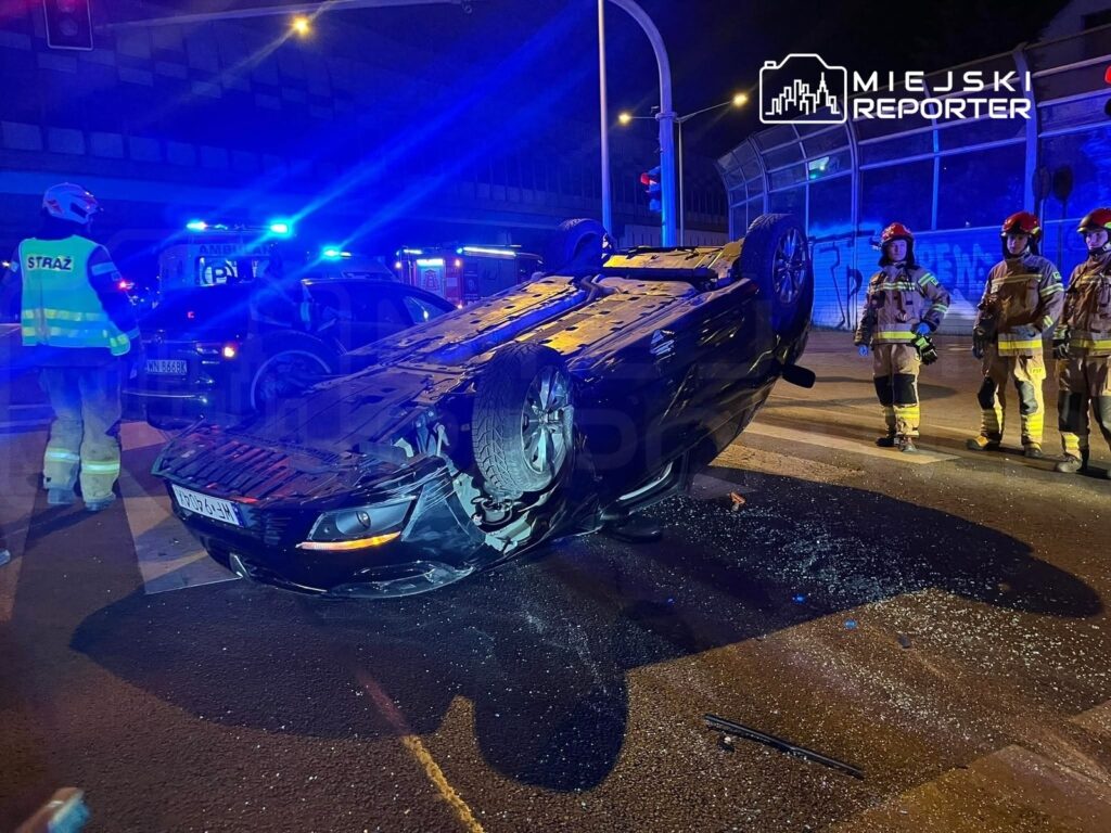 Czarny samochód leży na dachu na skrzyżowaniu, obok grupy strażaków w odblaskowych mundurach.