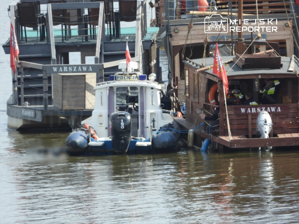 Policyjny statek motorowy zacumowany obok drewnianej łodzi wypełnionej ludźmi, w tle woda i flagi.