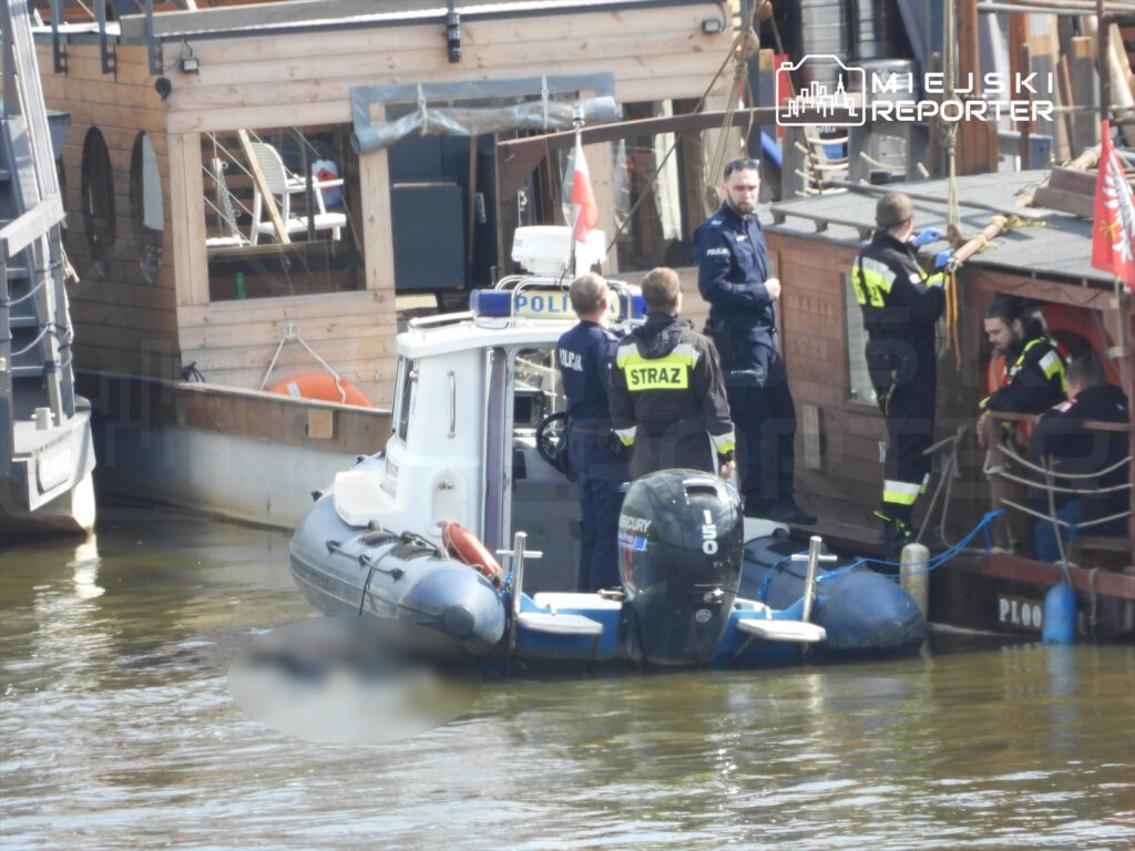 Grupa ratowników i policjantów na łodzi przy molo, prowadząca działania w pobliżu drewnianego statku.