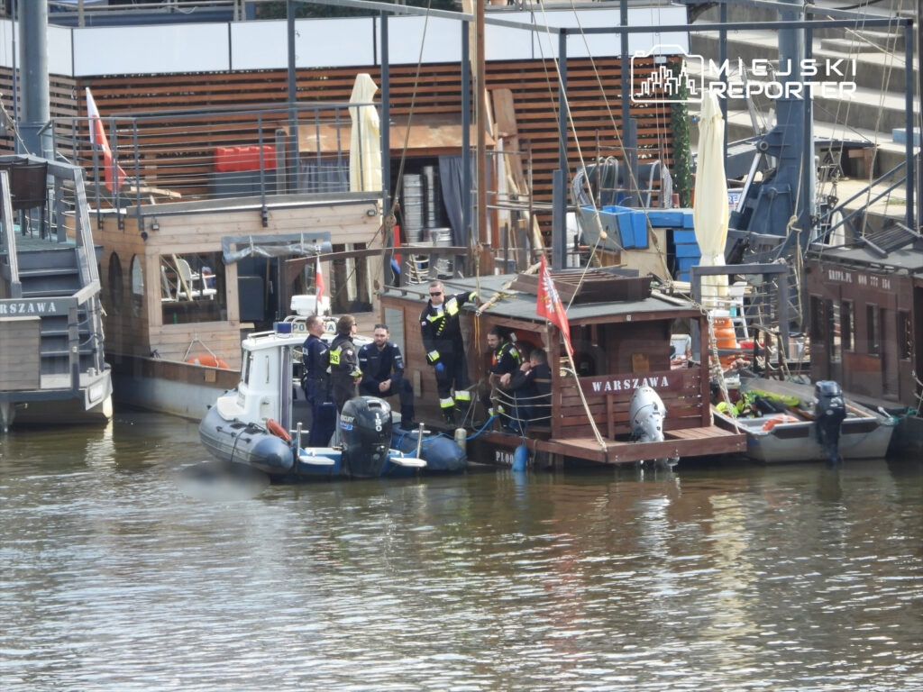 Grupa strażaków i policjantów rozmawia na pokładzie drewnianej łodzi przy nabrzeżu, obok małego kutra.
