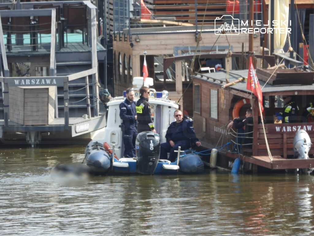 Dwóch policjantów na małej łodzi motorowej, zatrzymanych w porcie, obok drewnianych bark.