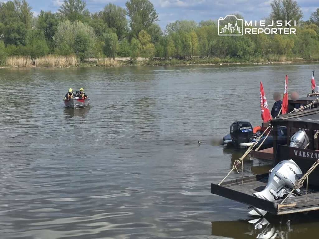 Dwóch ratowników w kamizelkach ochronnych płynie łodzią wzdłuż rzeki, w tle widoczne zarośla i drzewa.