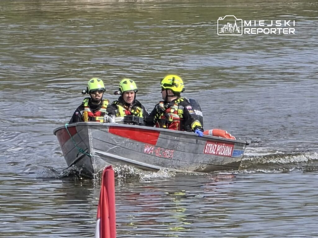 Trzech ratowników w kamizelkach asekuracyjnych płynie łodzią po wodzie, w tle widać spokojną taflę rzeki.