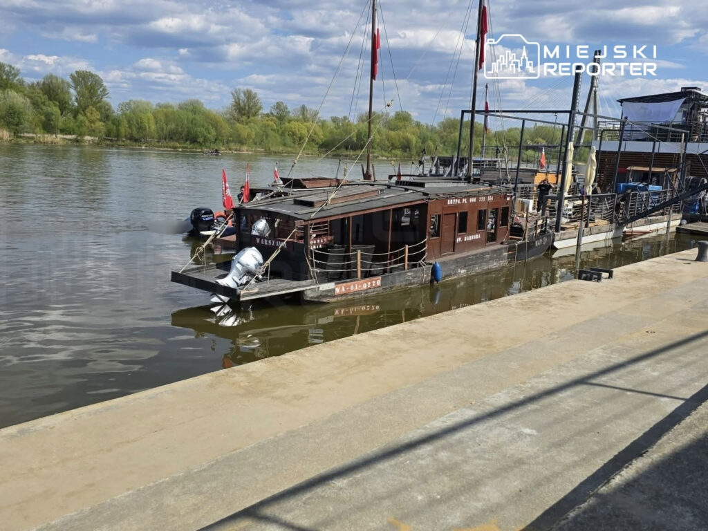 Statek z przeszkloną nadbudówką cumujący przy nabrzeżu, otoczony zielenią i wodą, z flagami powiewającymi na maszcie.