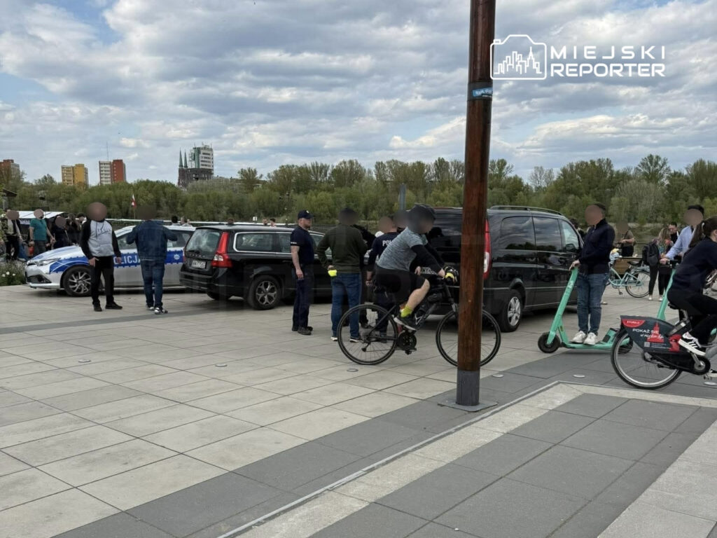 Grupa osób gromadzi się w pobliżu radiowozu policyjnego na placu, a niektórzy poruszają się na rowerach i hulajnogach.