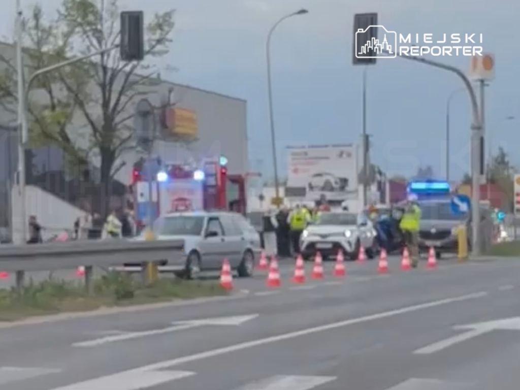 Grupa osób w odblaskowych kamizelkach zabezpiecza miejsce wypadku drogowego, obok stoją pojazdy służb ratunkowych.