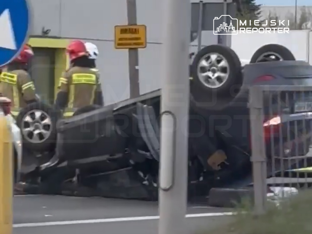 Strażacy w hełmach interweniują przy przewróconym samochodzie na drodze, obok znaku drogowym.