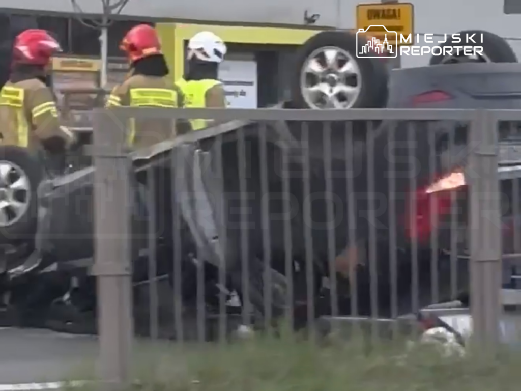 Strażacy w kaskach pomagają przy przewróconym samochodzie na ruchliwej drodze, w tle widoczny znak ostrzegawczy.