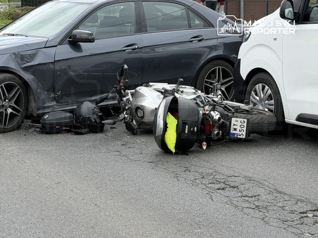 Motocykl leży na boku obok uszkodzonego samochodu osobowego na drodze. W tle widać drugi samochód.