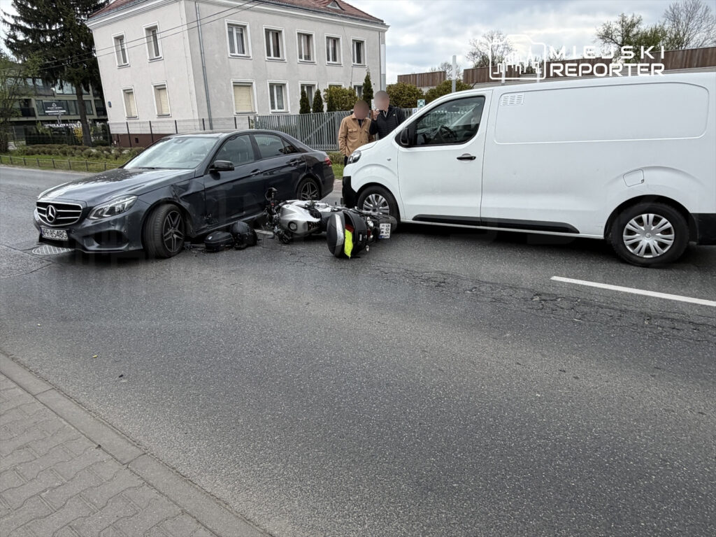 Na jezdni leży przewrócony motocykl obok dwóch uszkodzonych samochodów, w tym czarnego Mercedesa i białego furgonu.