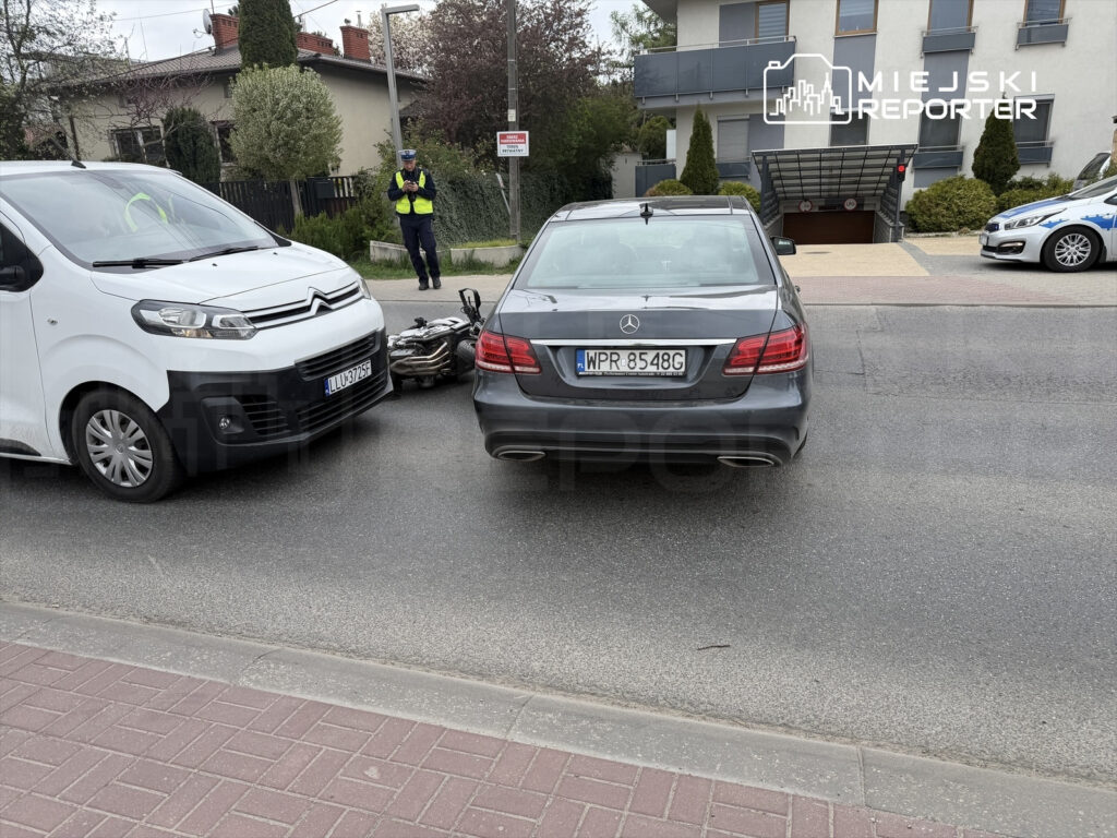 Kierowca białego vana zatrzymany obok leżącego motocykla na ulicy, w tle policjant dokumentujący zdarzenie.