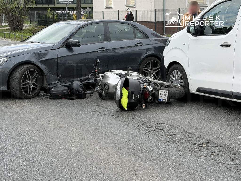 Motocykl leży na jezdni obok uszkodzonego samochodu osobowego, w tle widoczna inna furgonetka.