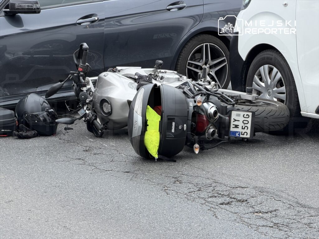 Motocykl leżący na boku na drodze, obok kask oraz torba, w tle dwa zaparkowane samochody.