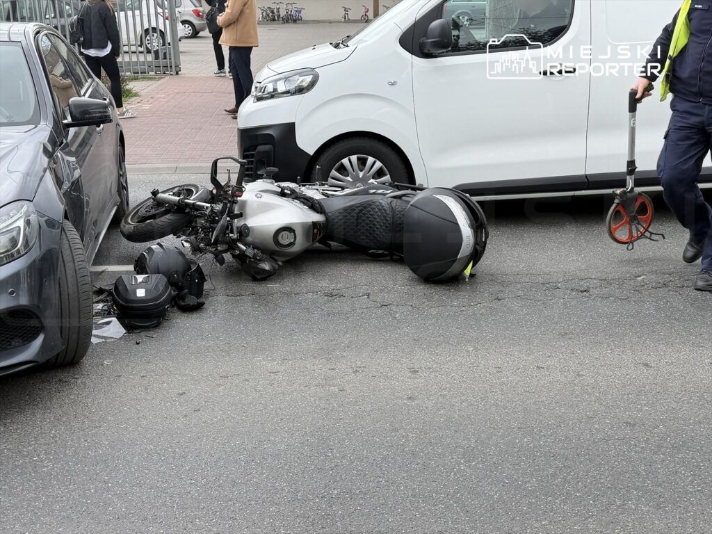 Motocykl leży na jezdni obok zaparkowanych samochodów, obok niego kask i narzędzie do pomiaru.
