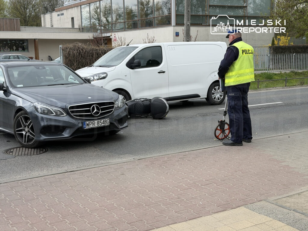 Policjant w żółtej kamizelce mierzy odległości na drodze obok uszkodzonego samochodu osobowego i dostawczego.