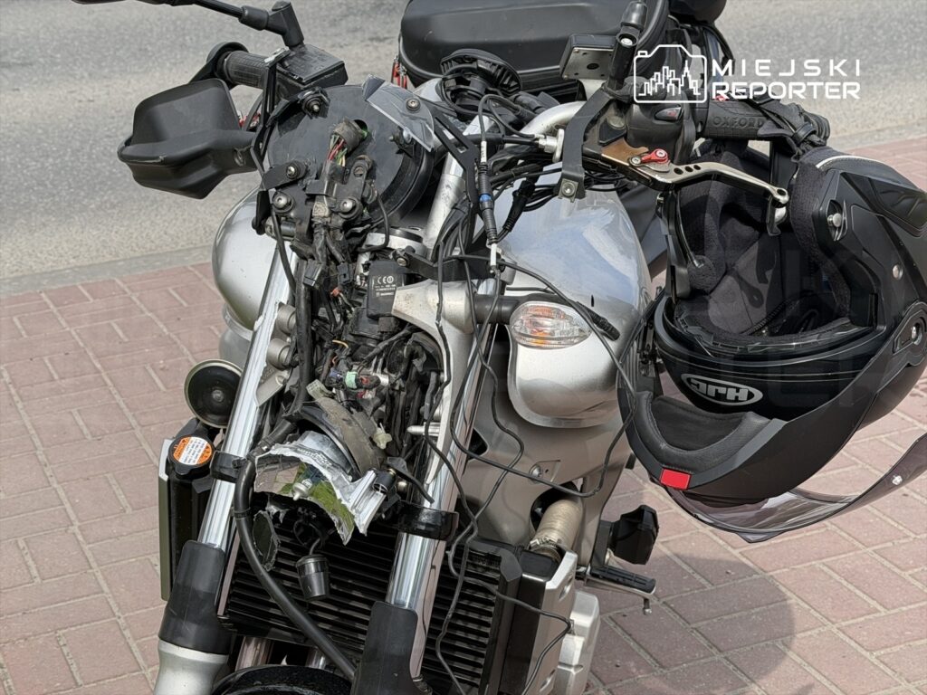 Motocykl z otwartą osłoną silnika, odsłaniającą przewody i podzespoły, stoi na chodniku obok kasku.