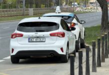 Biały Ford Focus zaparkowany obok taksówki na ulicy, z widoczną linią jezdni i chodnikiem.