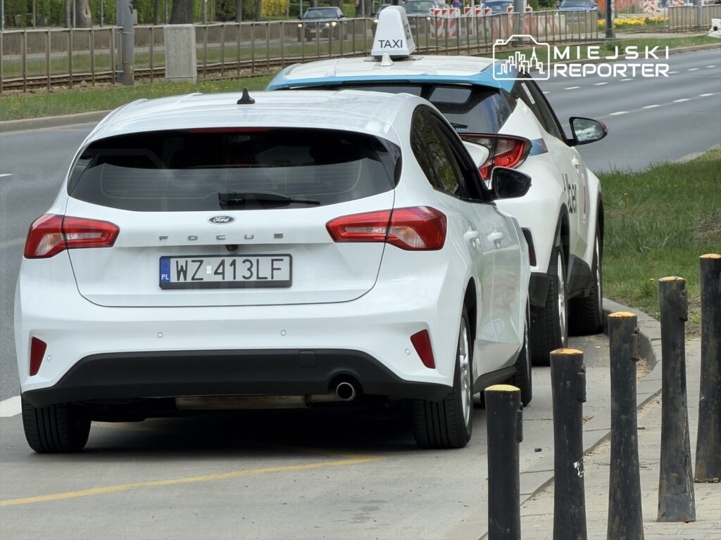 Biały Ford Focus zaparkowany obok taksówki z niebieskim dachem na miejskiej ulicy.