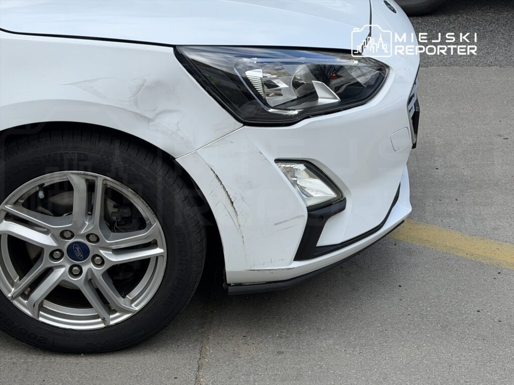 Biały samochód osobowy z uszkodzoną przednią częścią i zarysowaniami na nadwoziu stoi na parkingu.