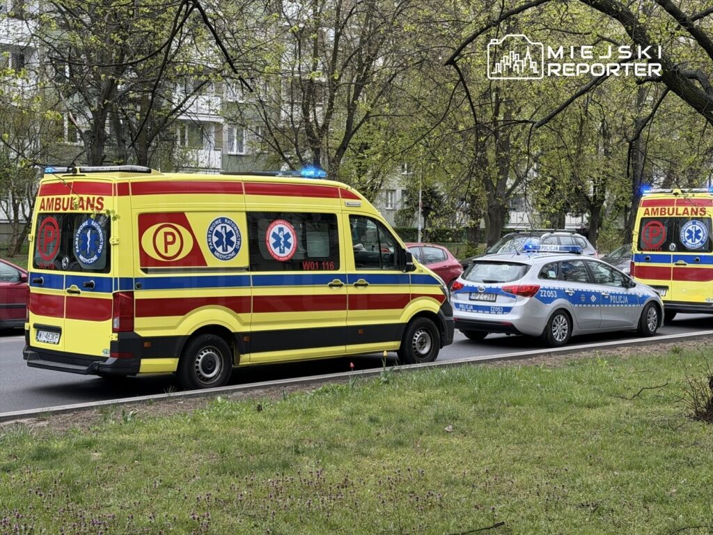 Karetka pogotowia oraz radiowóz policyjny zatrzymane na drodze, otoczone drzewami i budynkami mieszkalnymi.