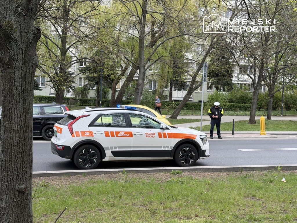 Radiowóz z niebieskim światłem zatrzymany na drodze, obok policjant kontrolujący ruch w miejskim otoczeniu.