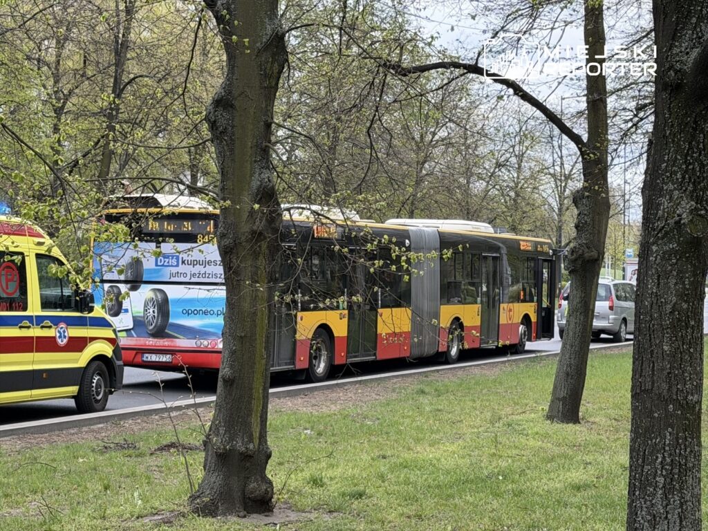 Karetka pogotowia i autobus miejski stoją na ulicy, otoczone drzewami, wiosenna zieleń w tle.