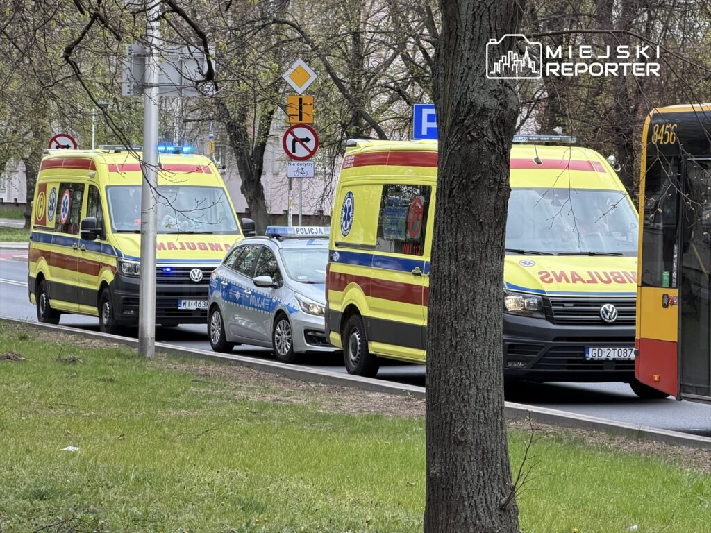 Dwa karetki pogotowia z włączonymi sygnałami świetlnymi stoją obok radiowozu policyjnego na miejskiej ulicy.