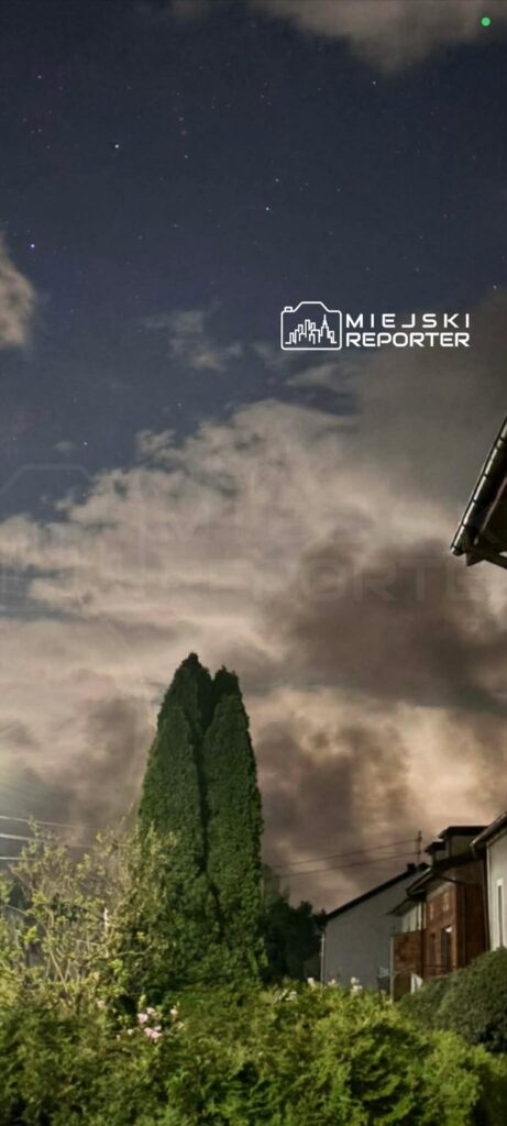 Nocne niebo z gwiazdami, chmury i wysokie drzewa w tle, w otoczeniu domów.