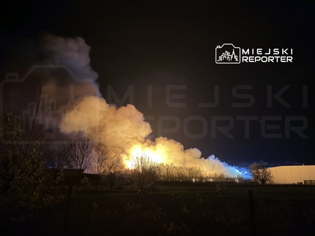Gęsty, pomarańczowy dym wydobywający się z pożaru w nocy, w tle widać kontury drzew i budynków.