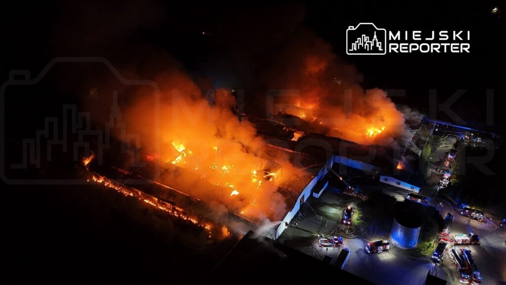 Pożar dużego budynku przemysłowego, z intensywnymi płomieniami i gęstym dymem, w otoczeniu wozów strażackich.