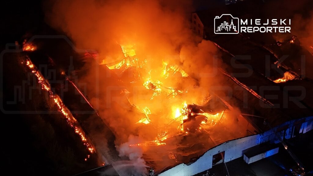 Płonący budynek przemysłowy wypełniony ogniem i dymem, z widocznymi płomieniami oraz zniszczoną strukturą dachu.