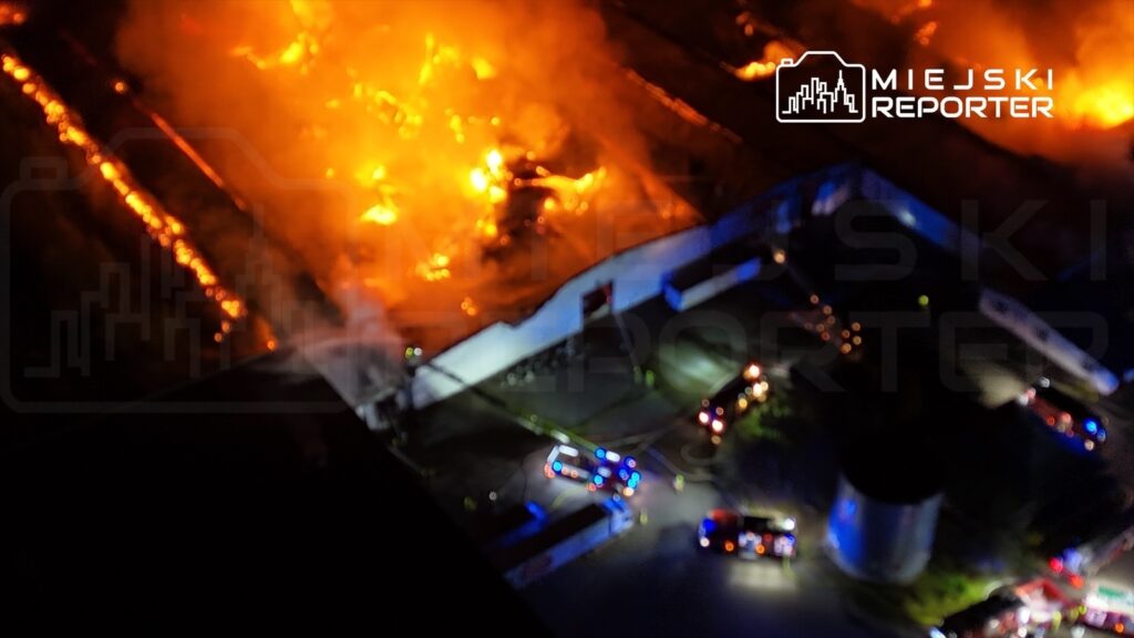 Intensywne płomienie i dym unoszący się nad budynkiem, przy którym pracują strażacy z wozami gaśniczymi.