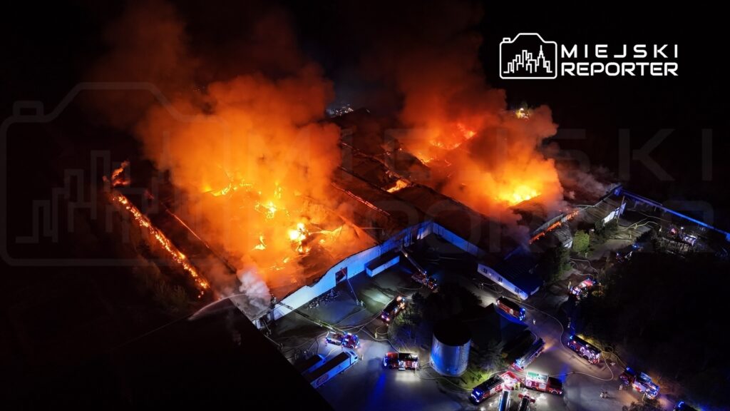 Ogromne płomienie wydobywające się z dachu magazynu, otoczone przez pojazdy straży pożarnej w nocy.