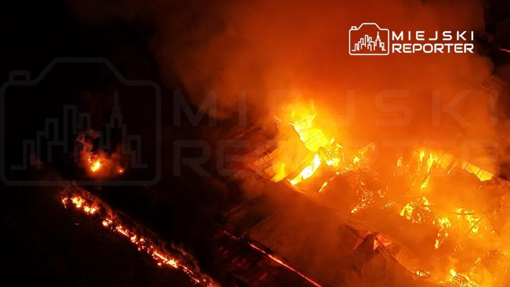 Wielki pożar trawi dach budynku, wydobywają się płomienie i dym w ciemnej scenerii nocnej.