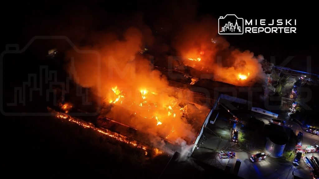Pożar dużego budynku przemysłowego, otoczony gęstym dymem i płomieniami, z wozami strażackimi w tle.