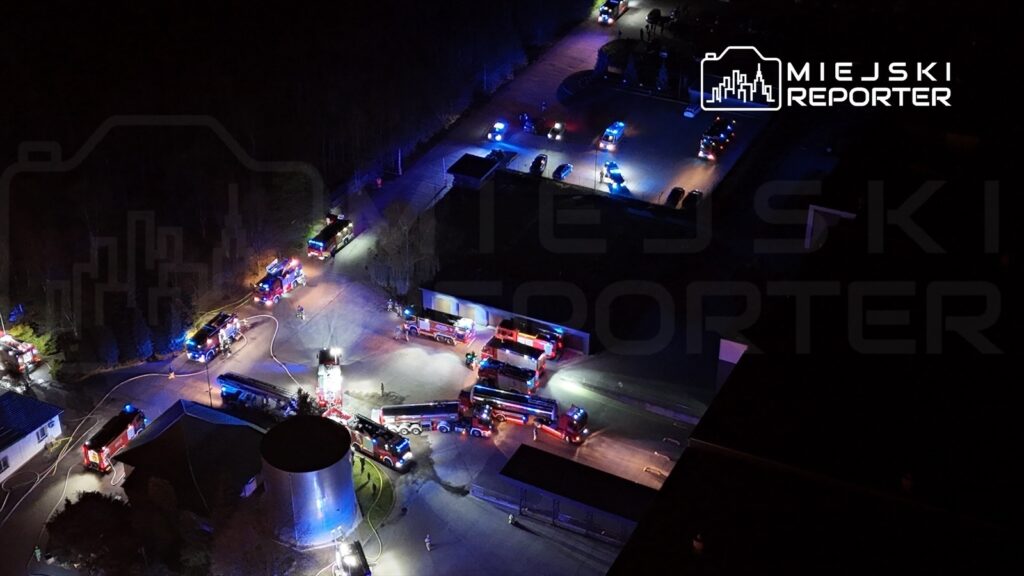 Grupa wozów strażackich z włączonymi sygnałami świetlnymi manewruje w okolicy budynku w nocy.