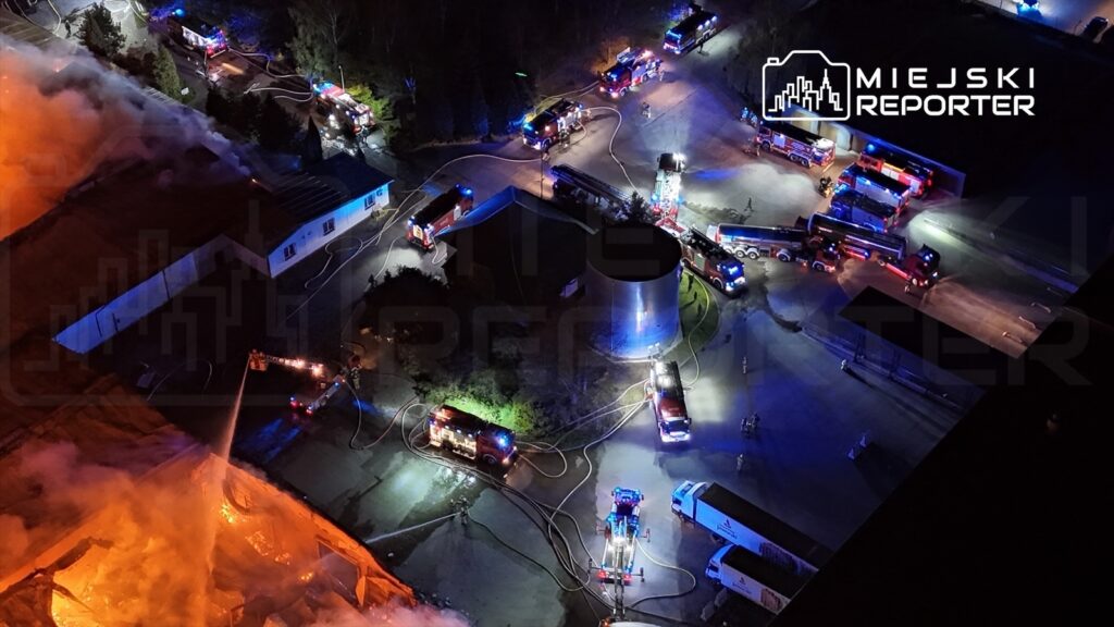 Strażacy walczą z pożarem w magazynie, otoczeni przez pojazdy gaśnicze i węże, w nocnym otoczeniu.