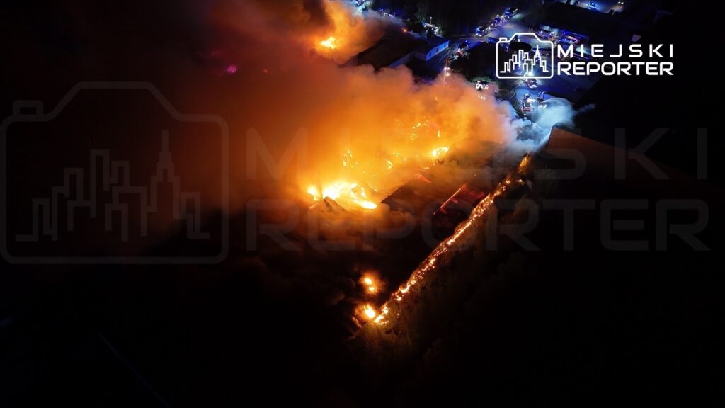Intensywny pożar z dużymi płomieniami i gęstym dymem w nocnym krajobrazie, otoczenie z zabudowaniami i pojazdami ratunkowymi.
