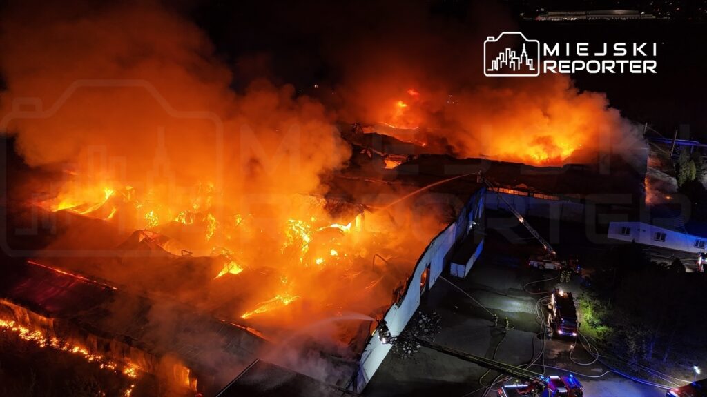Pożar trawi halę produkcyjną, a intensywne płomienie i dym unoszą się w nocy, podczas akcji gaśniczej.