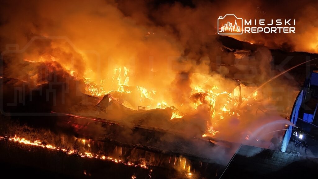 Wielka pożar na terenie przemysłowym, ogień trawi konstrukcję budynku, a w tle widoczne są strugi wody gaszące płomienie.